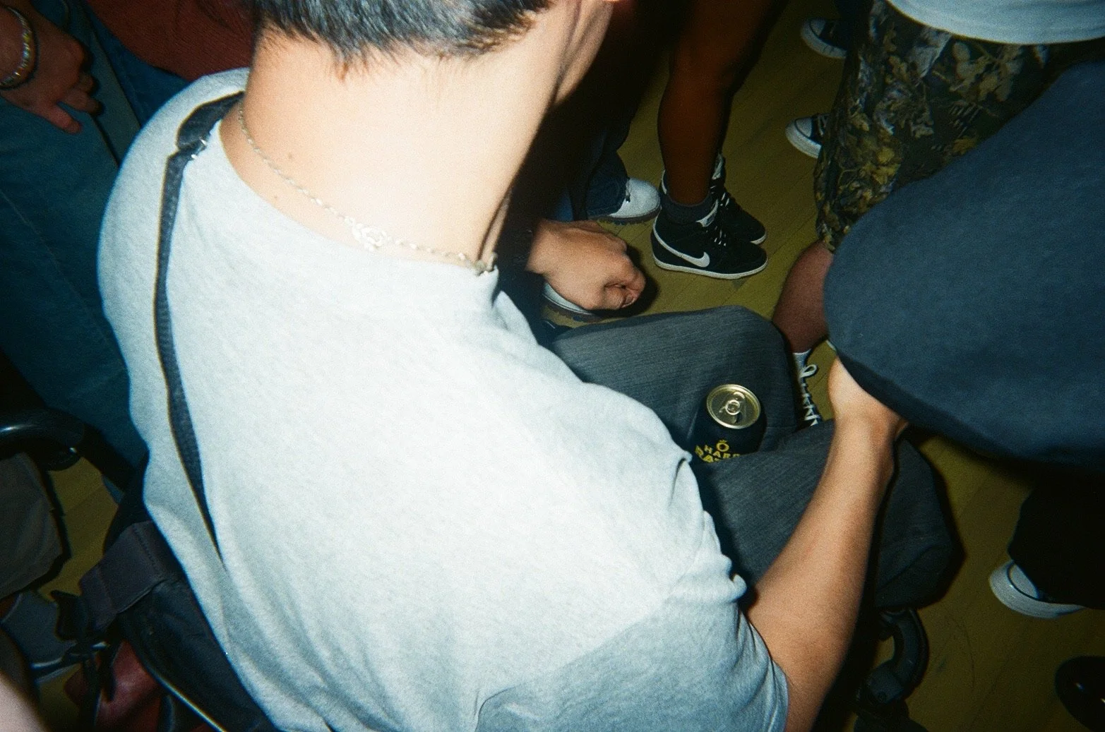 A person with short dark hair wearing a light gray T-shirt and a necklace, sitting in a crowded place, holding a black backpack with a yellow can of beer partly visible in their lap.