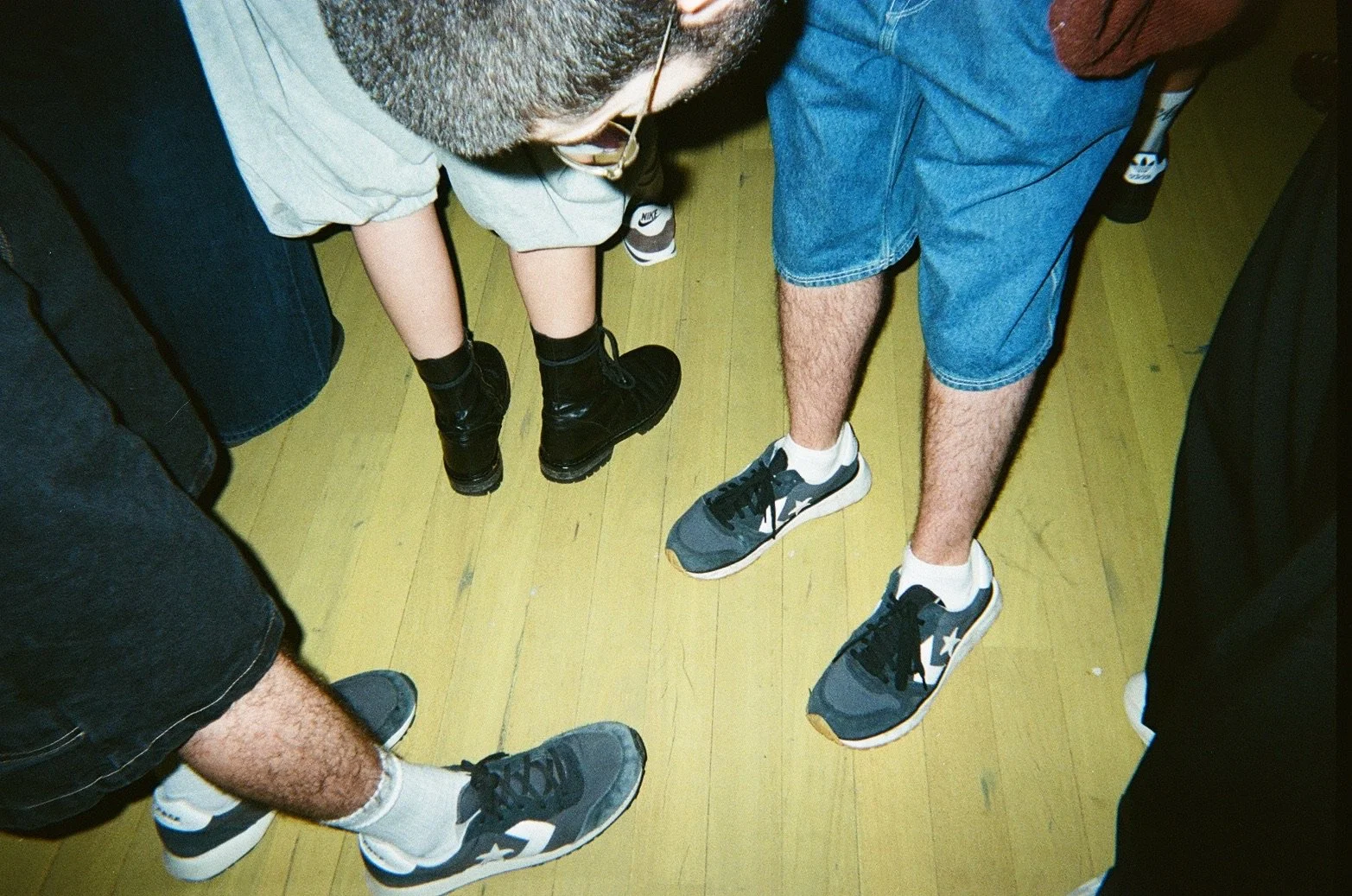 Top-down view of five people standing in a circle on a yellow wooden floor, showing their legs and feet. They are wearing sneakers and casual clothing.
