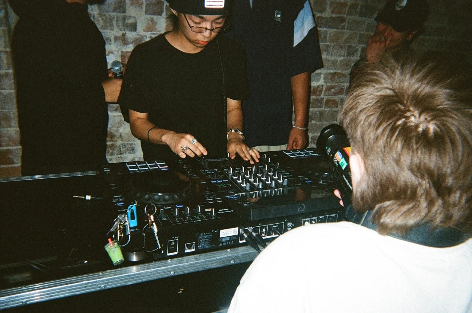 A woman DJ wearing glasses and jewelry is operating a DJ mixer and controller at a party or event with a brick wall background, with several people observing.