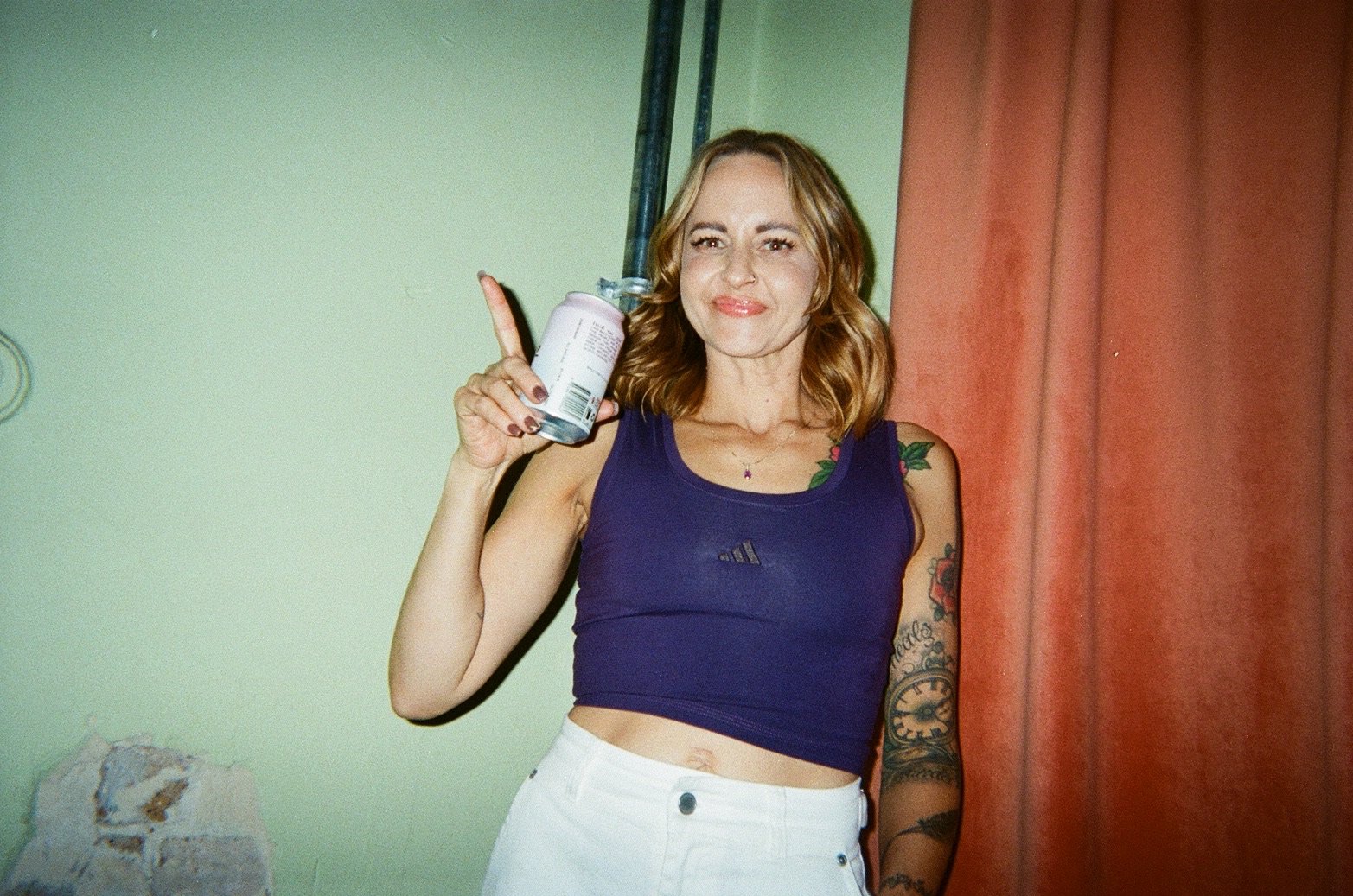 A woman with shoulder-length red hair, wearing a purple sleeveless top and white pants, standing against a light green wall and red curtain. She is holding a can of soda in her right hand and has tattoos on her left arm and shoulder.