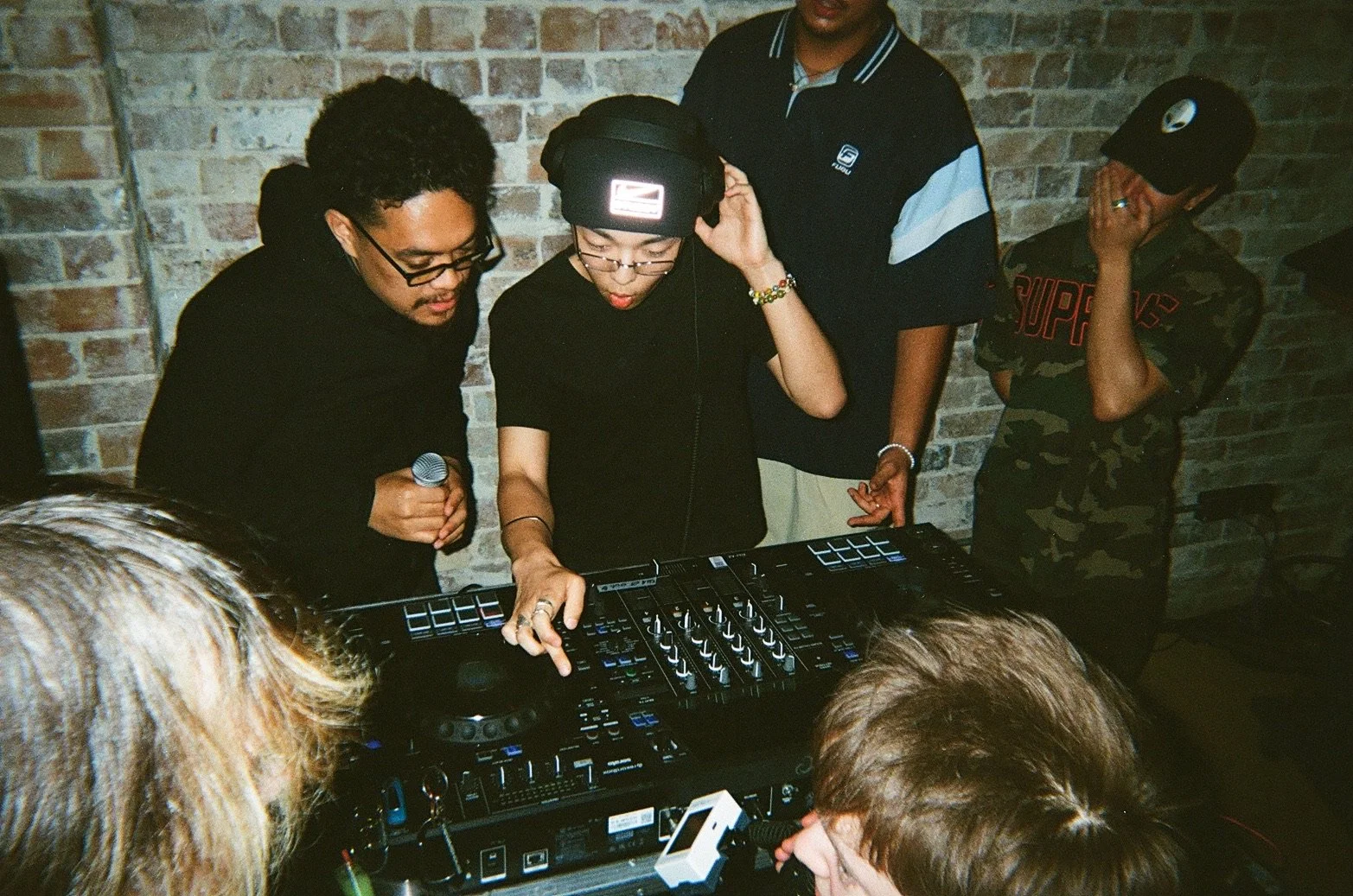 A group of people, including a DJ and an onlookers, gathered around a DJ controller in a room with exposed brick walls. The DJ is wearing glasses, a beanie, and is focused on the equipment, while others watch and listen.