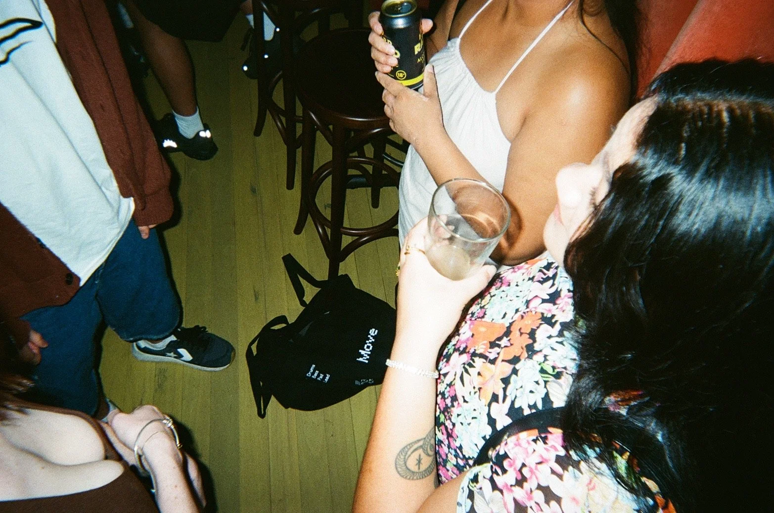 People socializing in a bar or restaurant, some holding drinks, with a wooden floor and stools in the background.
