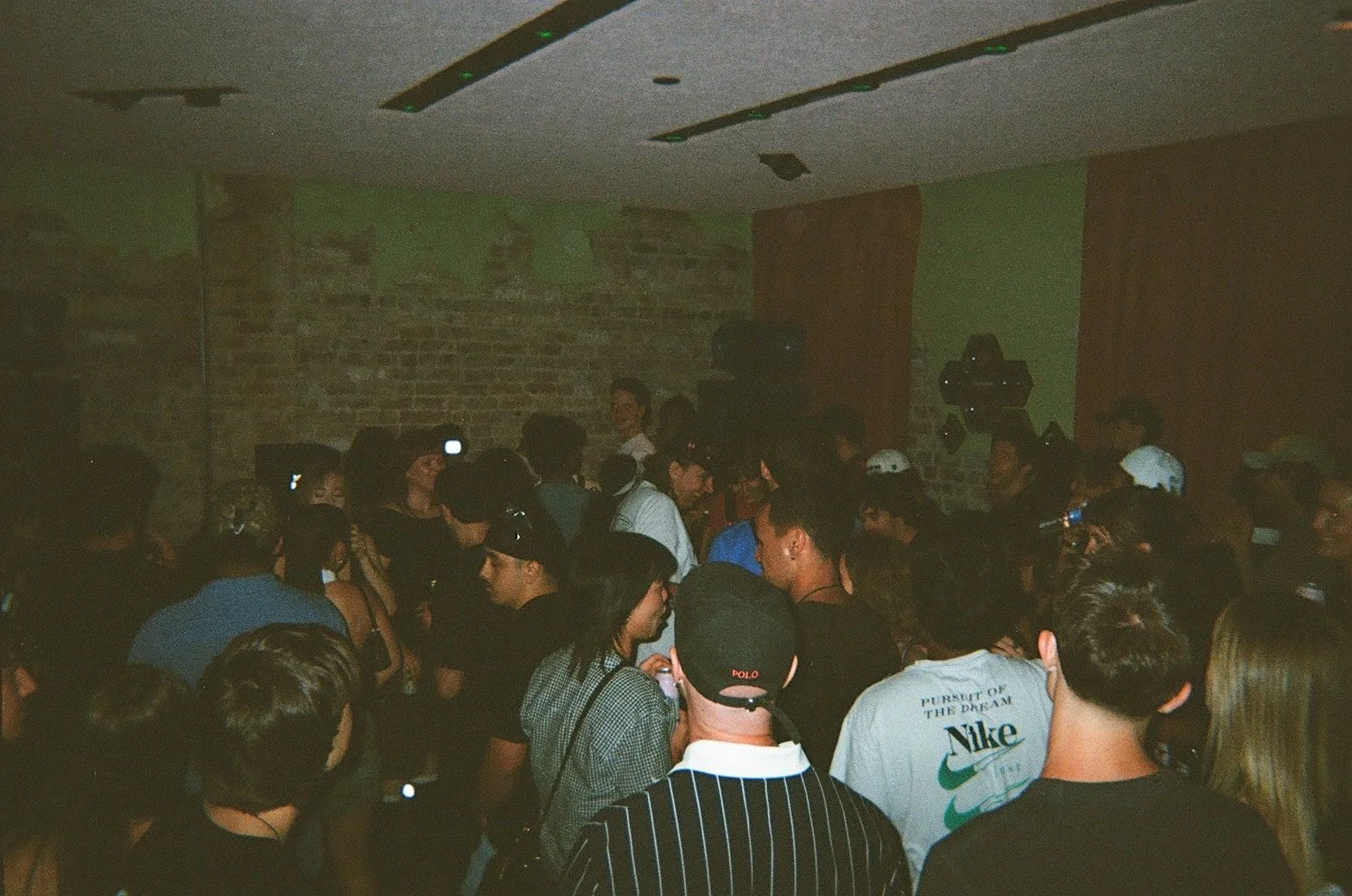 People gathered in a dimly lit indoor space with exposed brick walls and some speakers on the wall, likely at a concert or party.