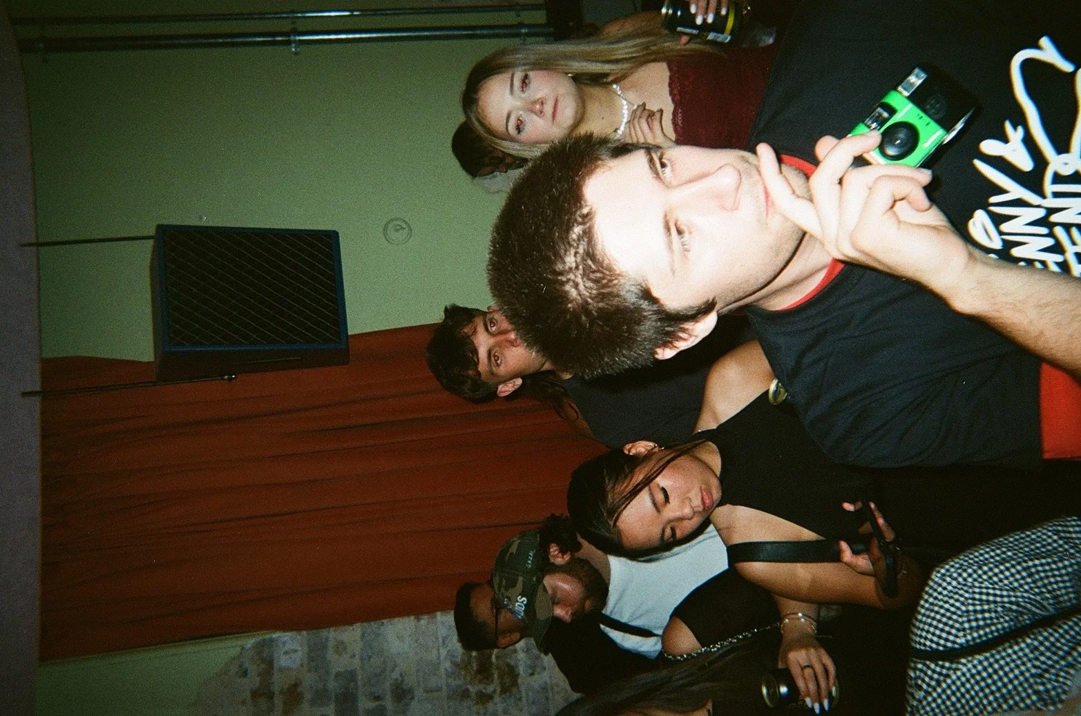 Group of people at a party, some holding drinks, standing near red curtains and a green wall with a black speaker, some looking at the camera, others looking away, casual attire.