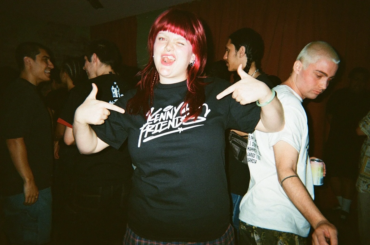 A young woman with red hair and a nose piercing making a silly face, pointing at herself with both hands. She is wearing a black T-shirt with the words 'KENNY FRIEND' on it. Behind her are other young people at a party or social gathering, some smili