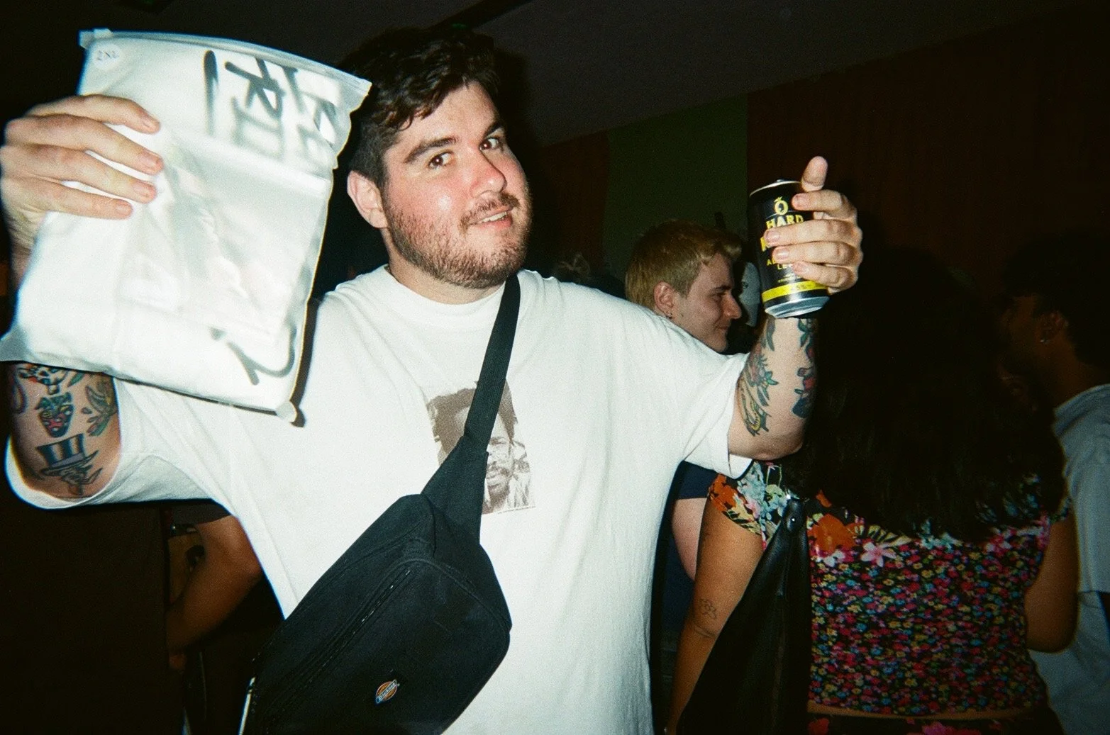 A man with dark hair and tattoos on his arms is holding a bag of chips in one hand and a can of beer in the other, smiling at the camera at a crowded indoor event.