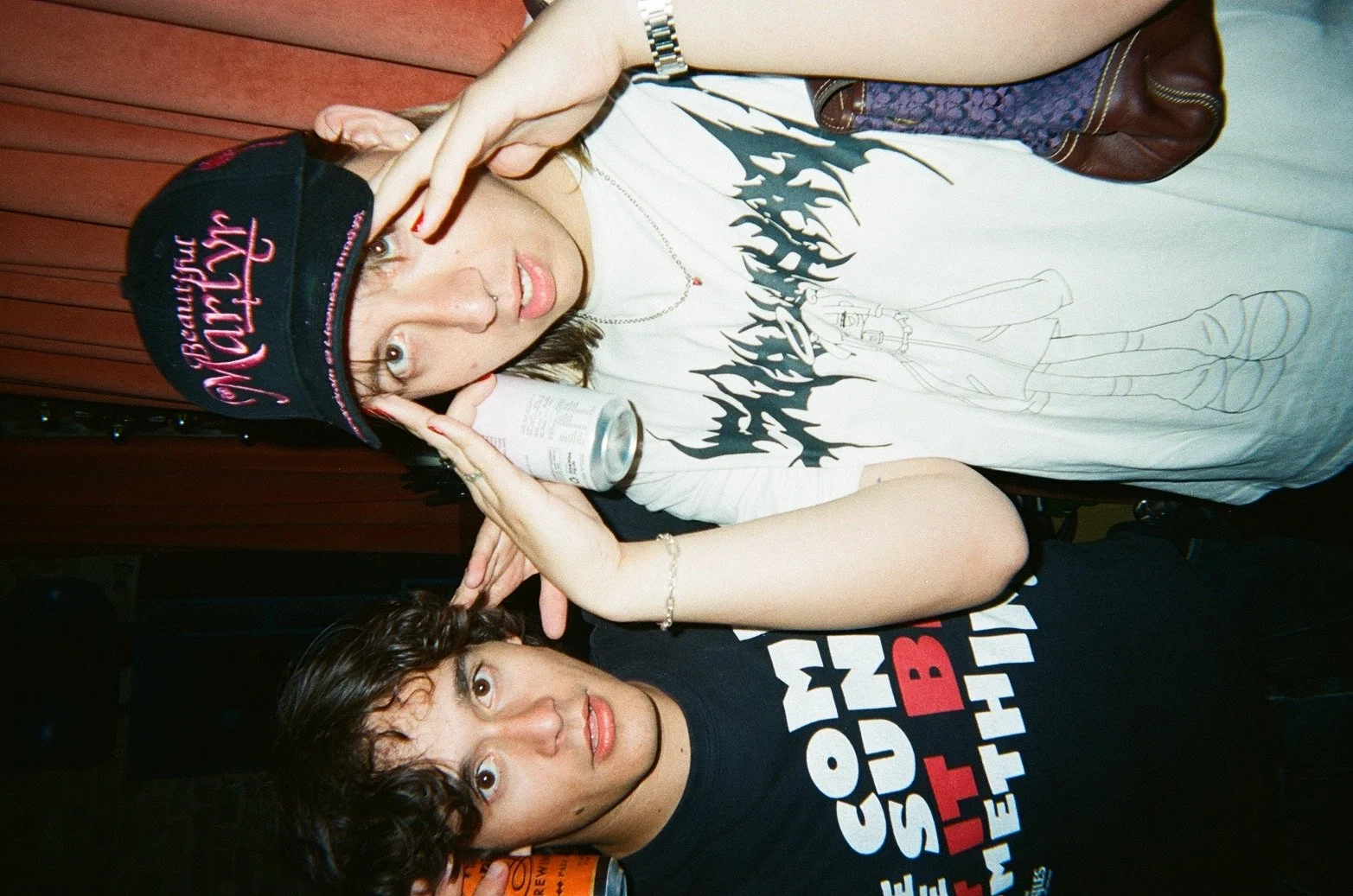 A woman wearing a black cap with pink writing, a necklace, and a graphic t-shirt making a gesture with her hand. Next to her is a man with curly hair wearing a black t-shirt with colorful text. Both appear to be at a social event.