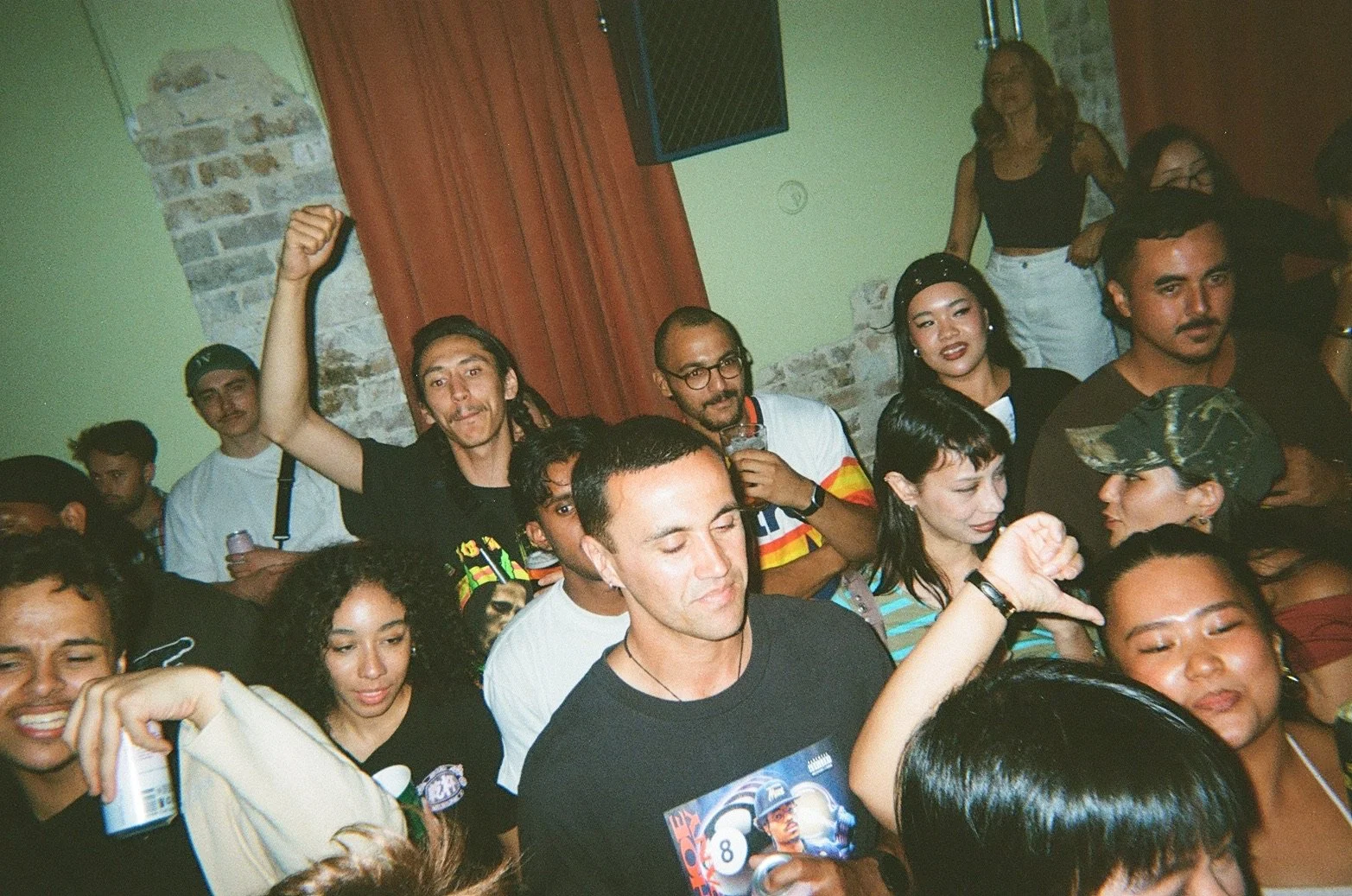 A diverse group of people at a social gathering or party, some dancing and enjoying drinks in a room with brick and green walls and red curtains.