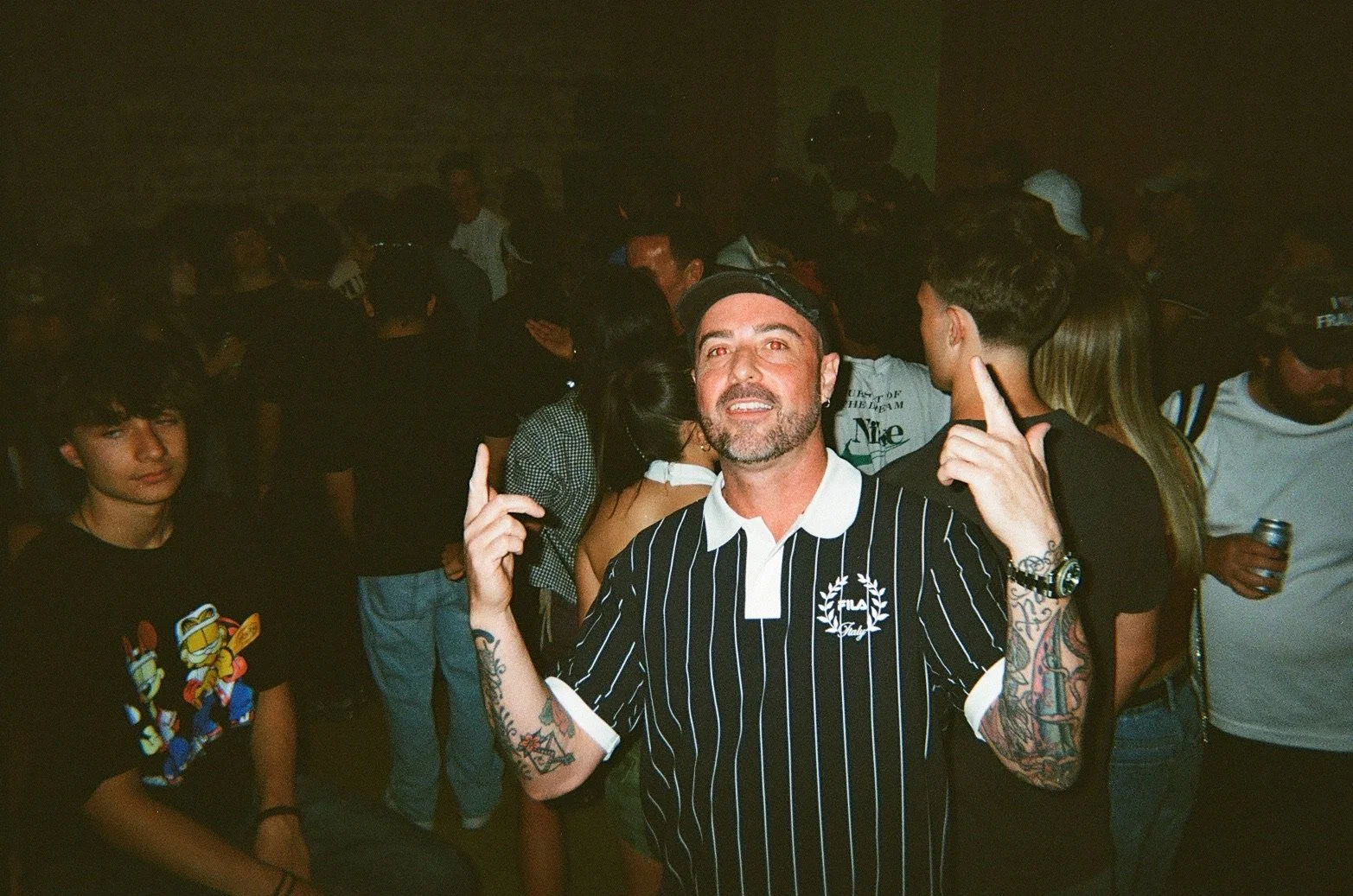 A man with tattoos on his arms, a beard, and wearing a striped black and white shirt, smiling and pointing upwards at a crowded party.