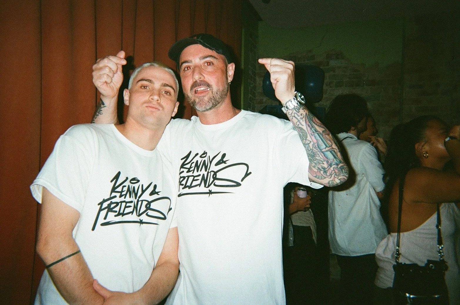 Two men posing with arm around each other at a social event, wearing white T-shirts with 'Lenny & Friends' logo, people in background, brick wall and wooden paneling.
