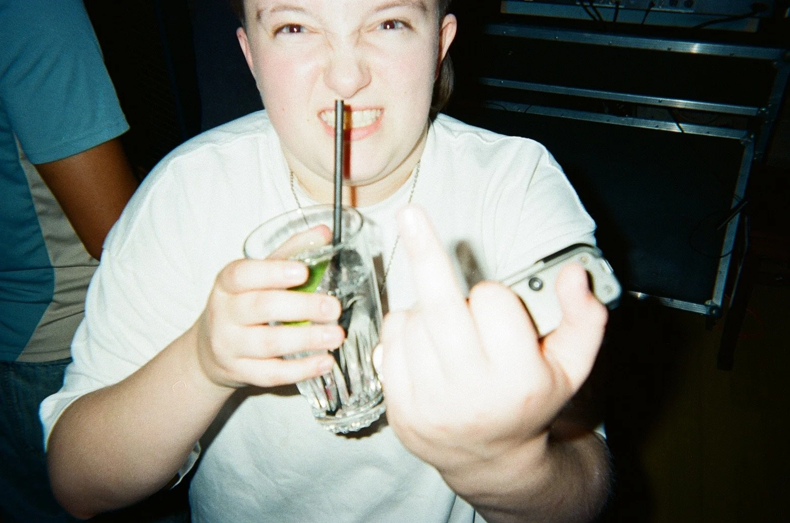 A young woman with dark hair wearing a white shirt, holding a drink and a phone, taking a selfie, with a playful expression showing teeth, at a social gathering or party.