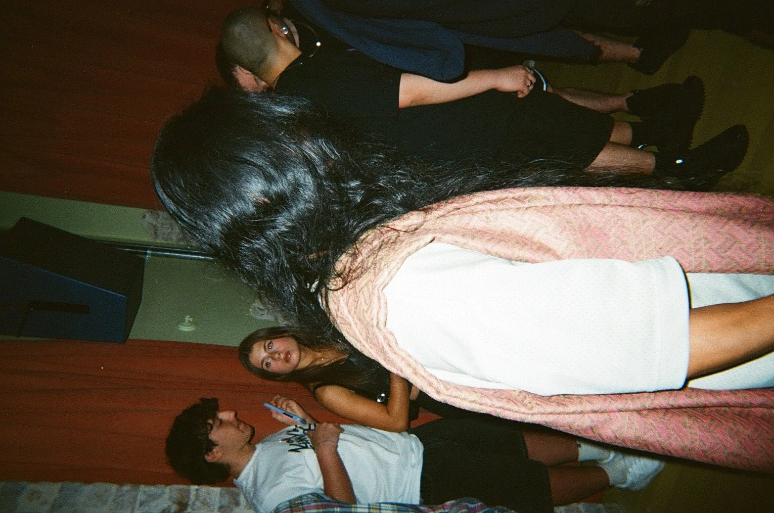 Two women are in the foreground, one with long black hair, wearing a white dress and a pink shawl, and another with long curly hair, wearing a beige dress. In the background, two men are standing, one with short hair and a black shirt, and another wi