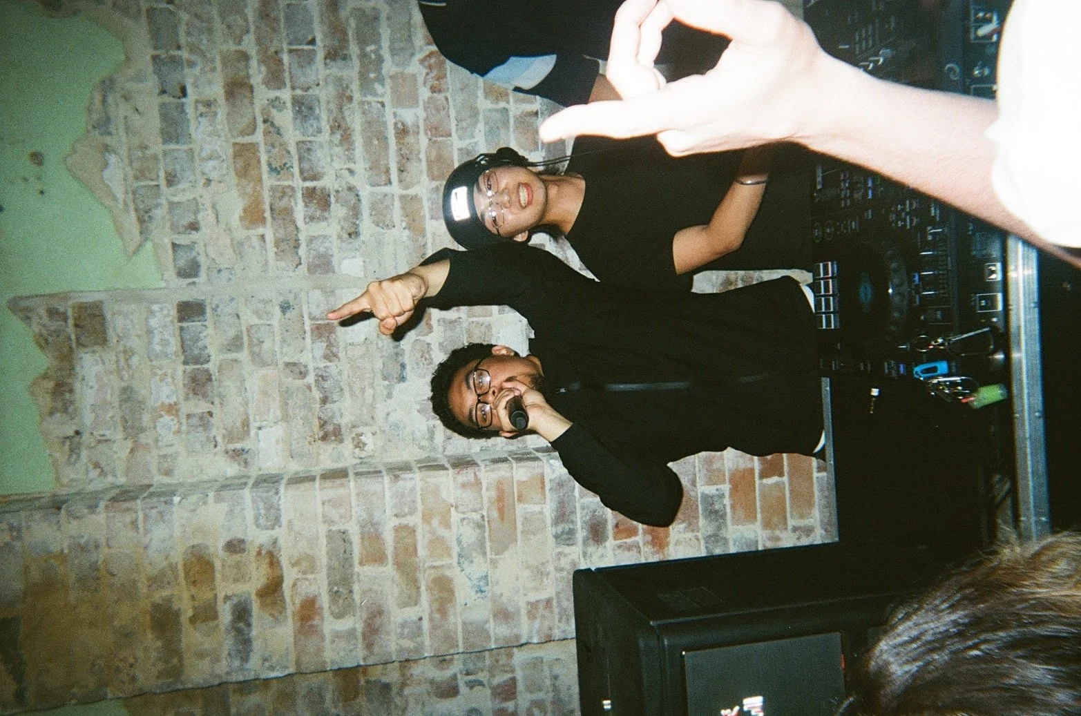 Two DJs in black outfits performing with a brick wall background, one wearing a cap and glasses, holding a microphone, the other making a gesture with a hand, surrounded by DJ equipment.