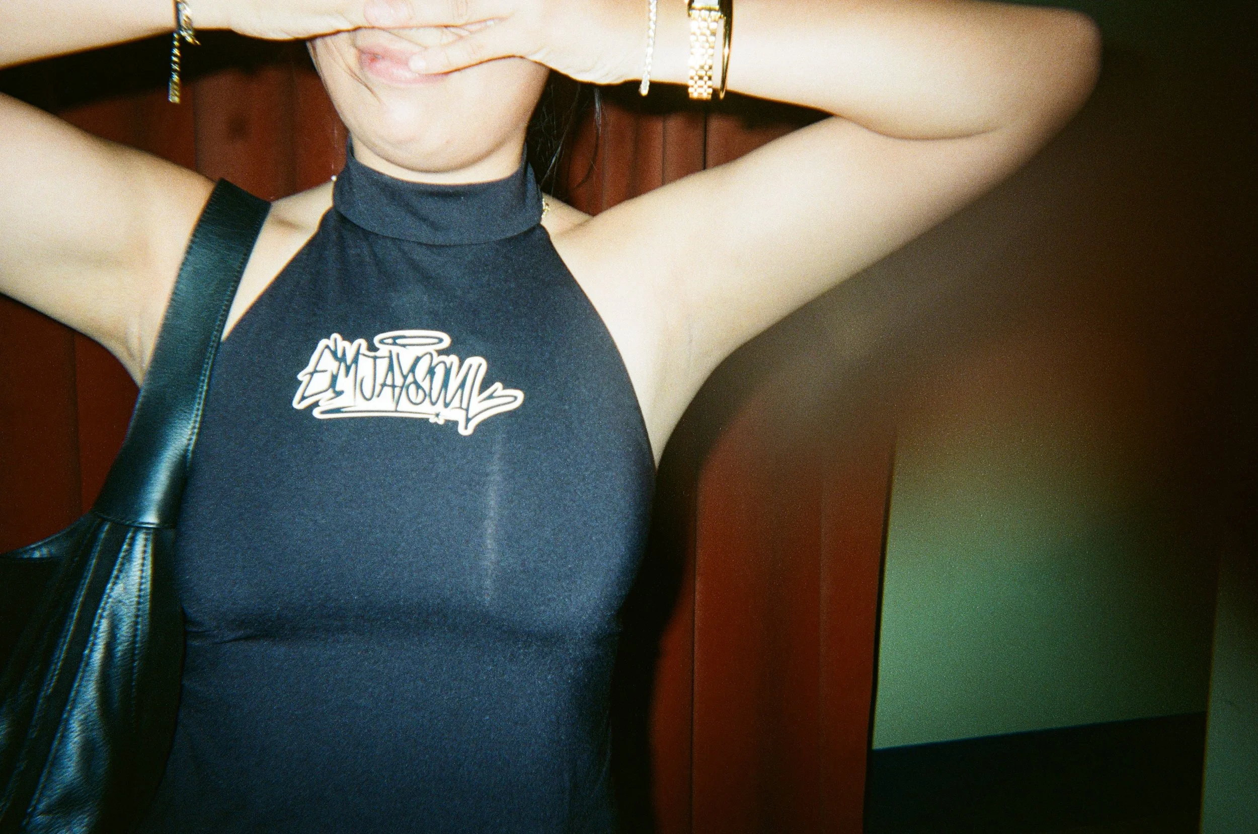 A woman wearing a black sleeveless top with the word 'AMAZON' printed on it, holding her hands up near her face, and carrying a black purse.