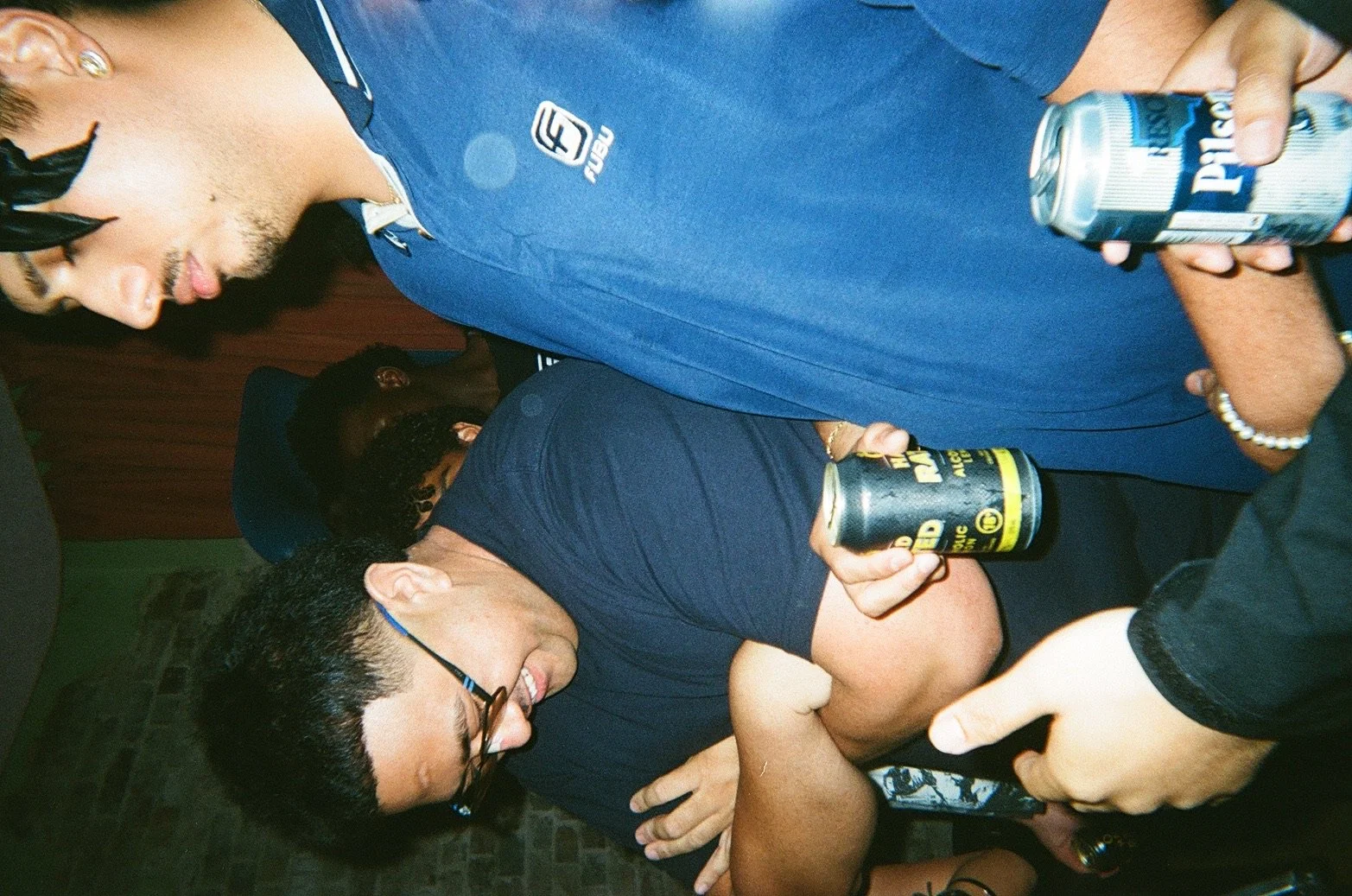 People dancing closely at a party, with some holding cans of beer and one person using a smartphone.