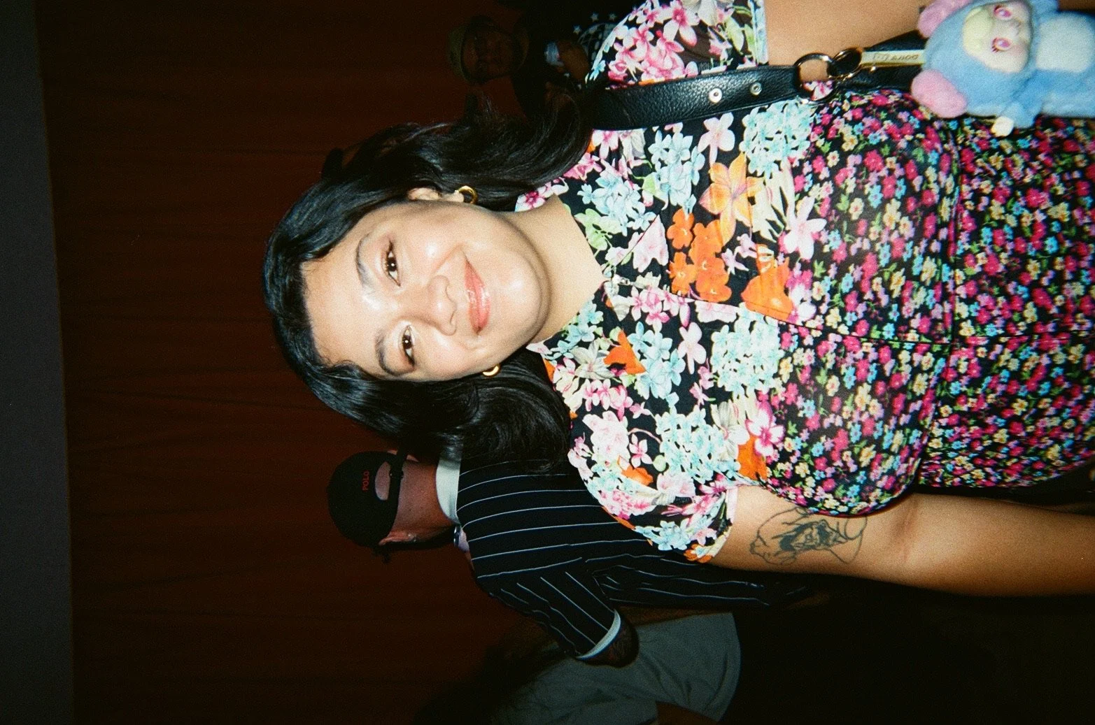 A woman with dark hair wearing a colorful floral dress poses for a photo. She is smiling and wearing gold earrings and a necklace with a small pendant. A person in a black striped shirt and cap stands behind her.