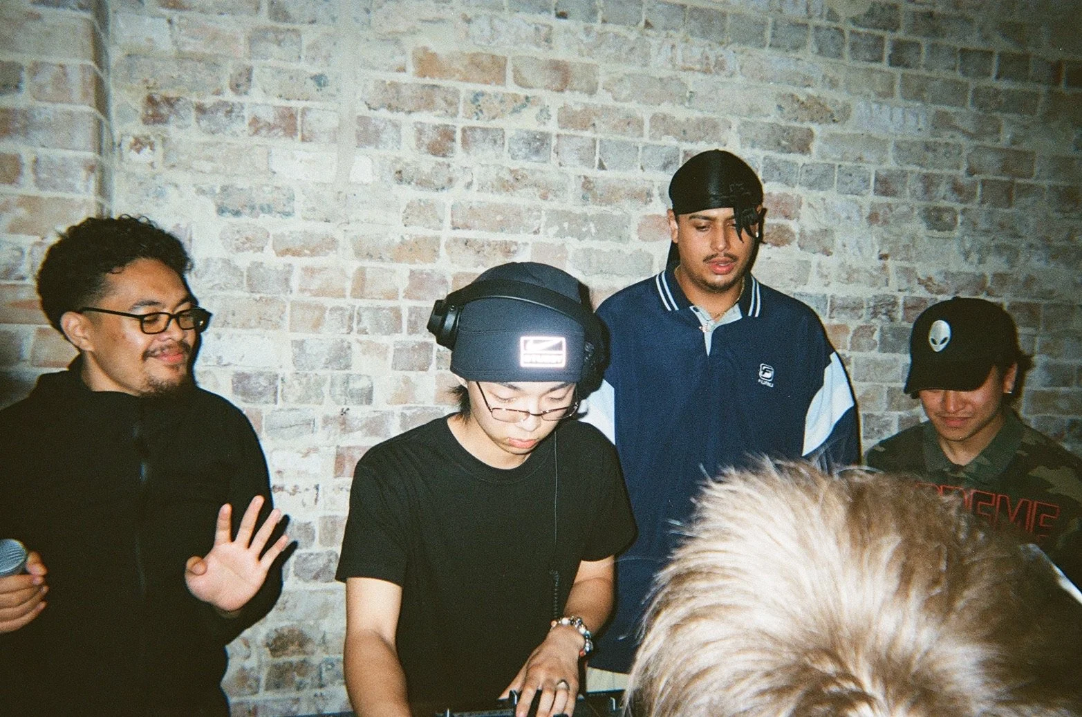 Group of five diverse people standing against a brick wall, with one person wearing headphones and a hat, and another with glasses, appearing to enjoy a music or DJ session.