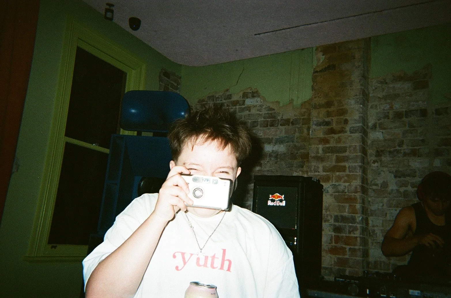 A person with short dark hair smiling and holding a camera up to their face, wearing a white t-shirt with the word 'yuth' on it, in a room with brick walls and a Red Bull vending machine in the background.