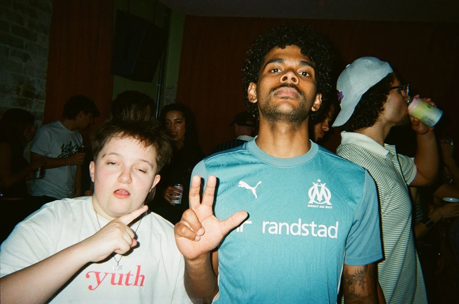 A young man in a light blue soccer jersey making a peace sign, standing next to a girl with short hair, a white shirt, and a necklace, who is also making a peace sign. They are at a social event with other people in the background.