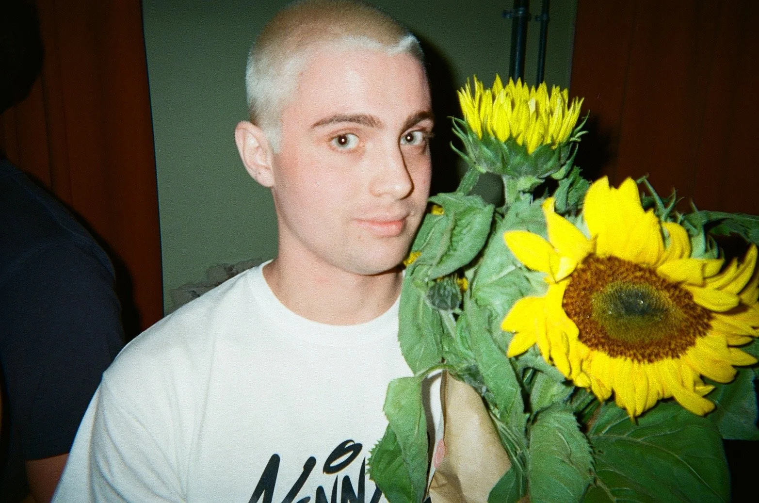 Person with very short blonde hair holding a bouquet of sunflowers.