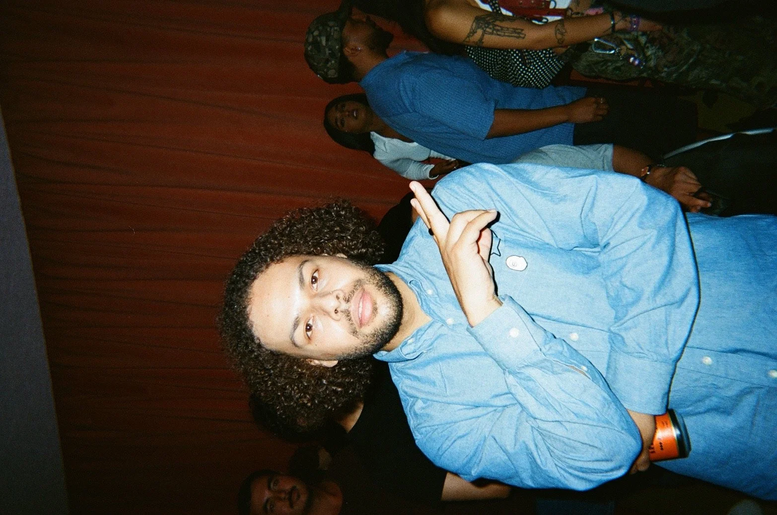 A young man with curly hair, a beard, and light skin, wearing a blue shirt, making a hand gesture with his fingers crossed, standing in front of a red curtain at an indoor event, with other people visible in the background.