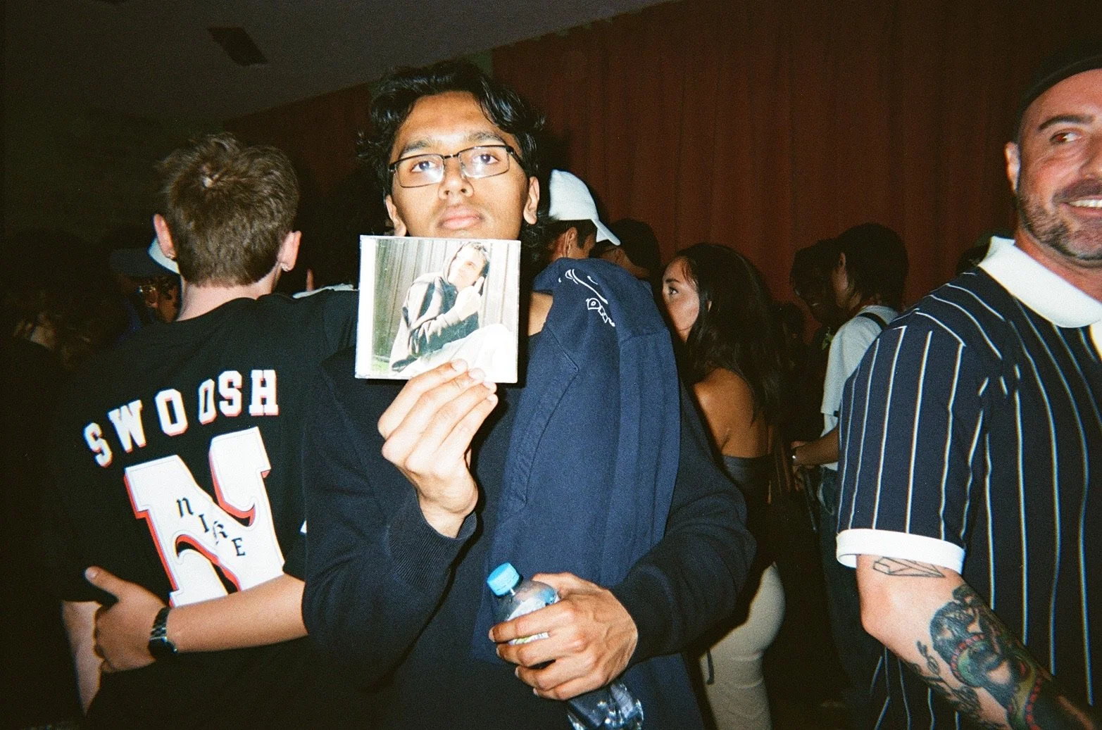A young man with glasses holding a photo of himself at an indoor event, surrounded by people including a person in a striped shirt with tattoos on their arm and someone in a black shirt with 'SNOW SH' written on the back.