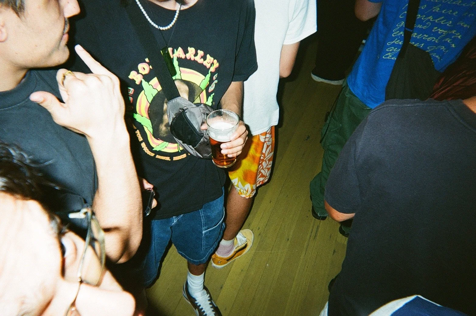 A group of people gathered at a party or social event. One person is holding a glass of beer and wearing a black t-shirt with colorful graphics. Others are dressed casually in t-shirts and shorts, and some are wearing glasses, necklaces, and watches.
