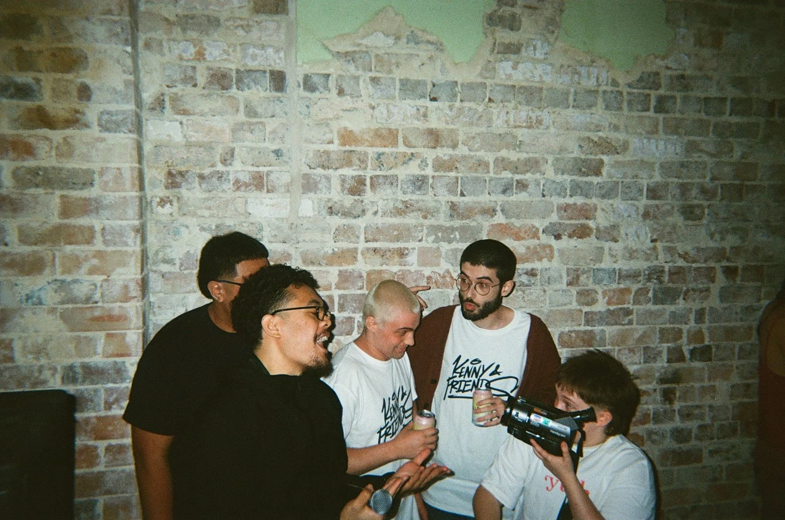 Group of five people gathered in front of a brick wall, one person is holding a video camera, others are talking and smiling