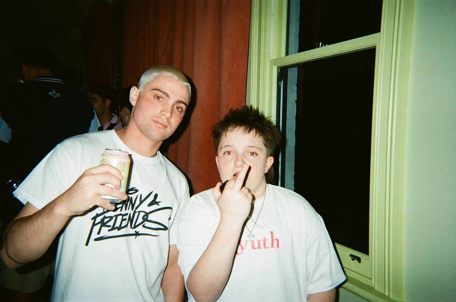 Two young men at a social event, one with bleached blonde hair and holding a drink, the other with short brown hair making a rude gesture near his face. They are standing indoors near a window with green trim, dark background with other people in the