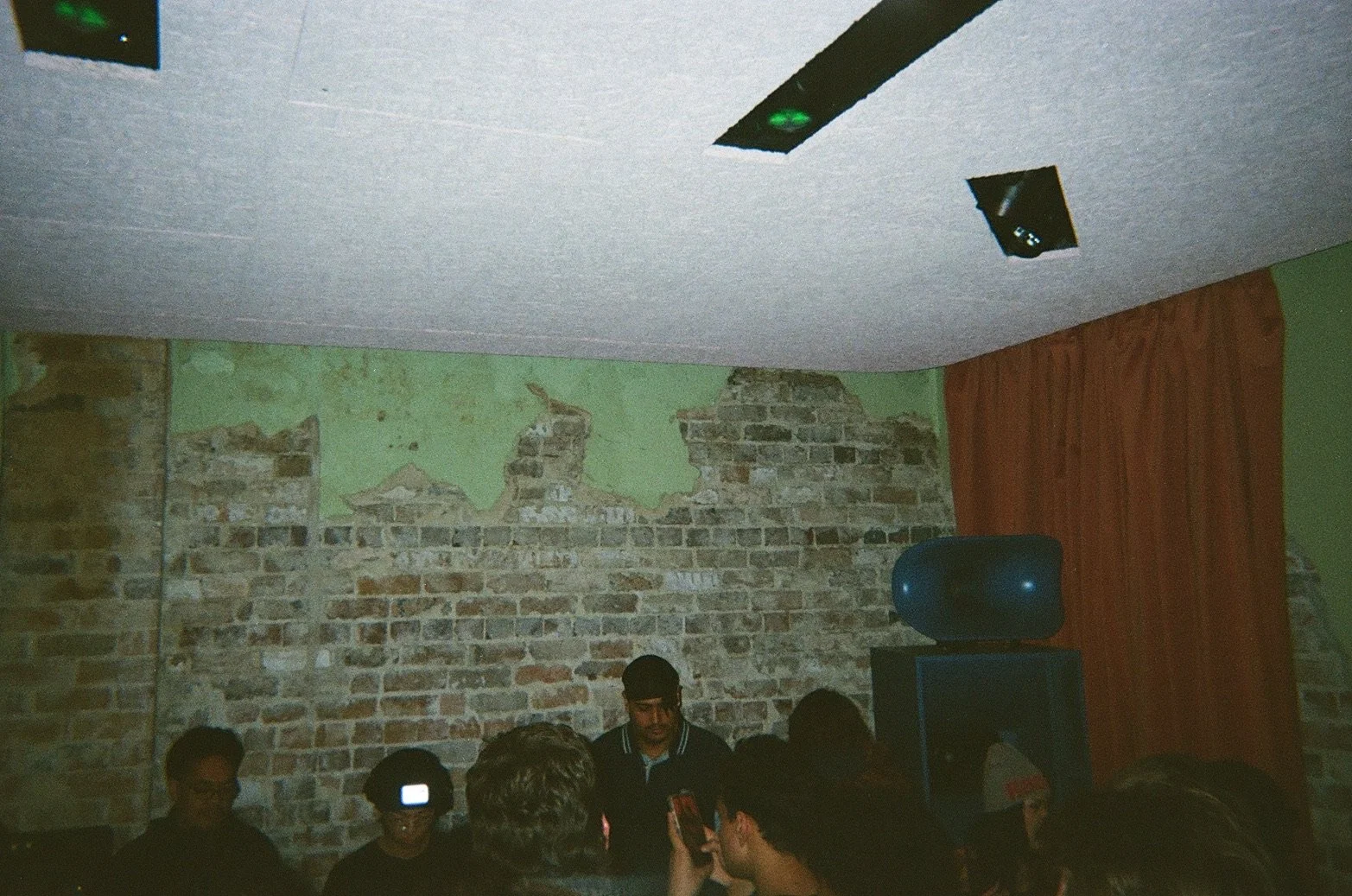 Group of people gathered in a room with exposed brick wall and red curtains, a man appears to be speaking or performing, with a satellite speaker and ceiling lights visible.