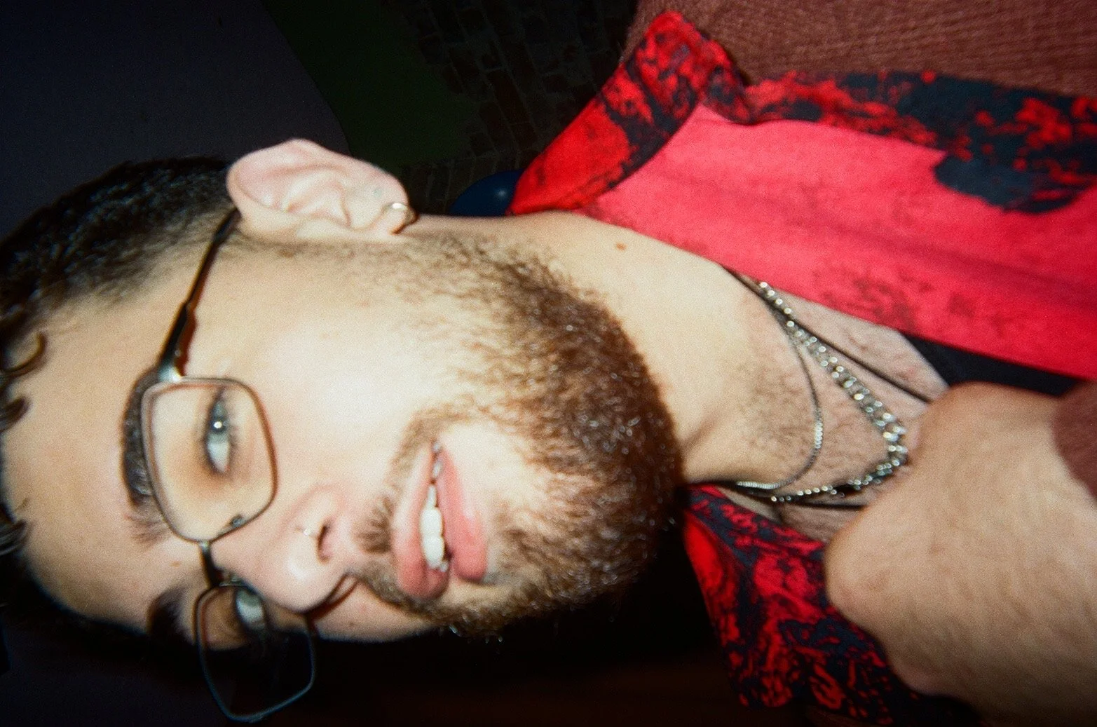 A young man with glasses, facial piercings, a beard, and a mustache is lying down, wearing a silver chain necklace and a red and black patterned shirt. The photo is taken at a close angle from above.