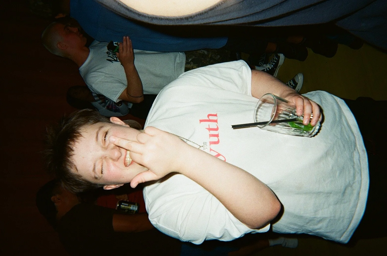 A young man at a party or social gathering making a funny face, pinching his nose, with a drink in his hand, standing next to a group of people in the background.