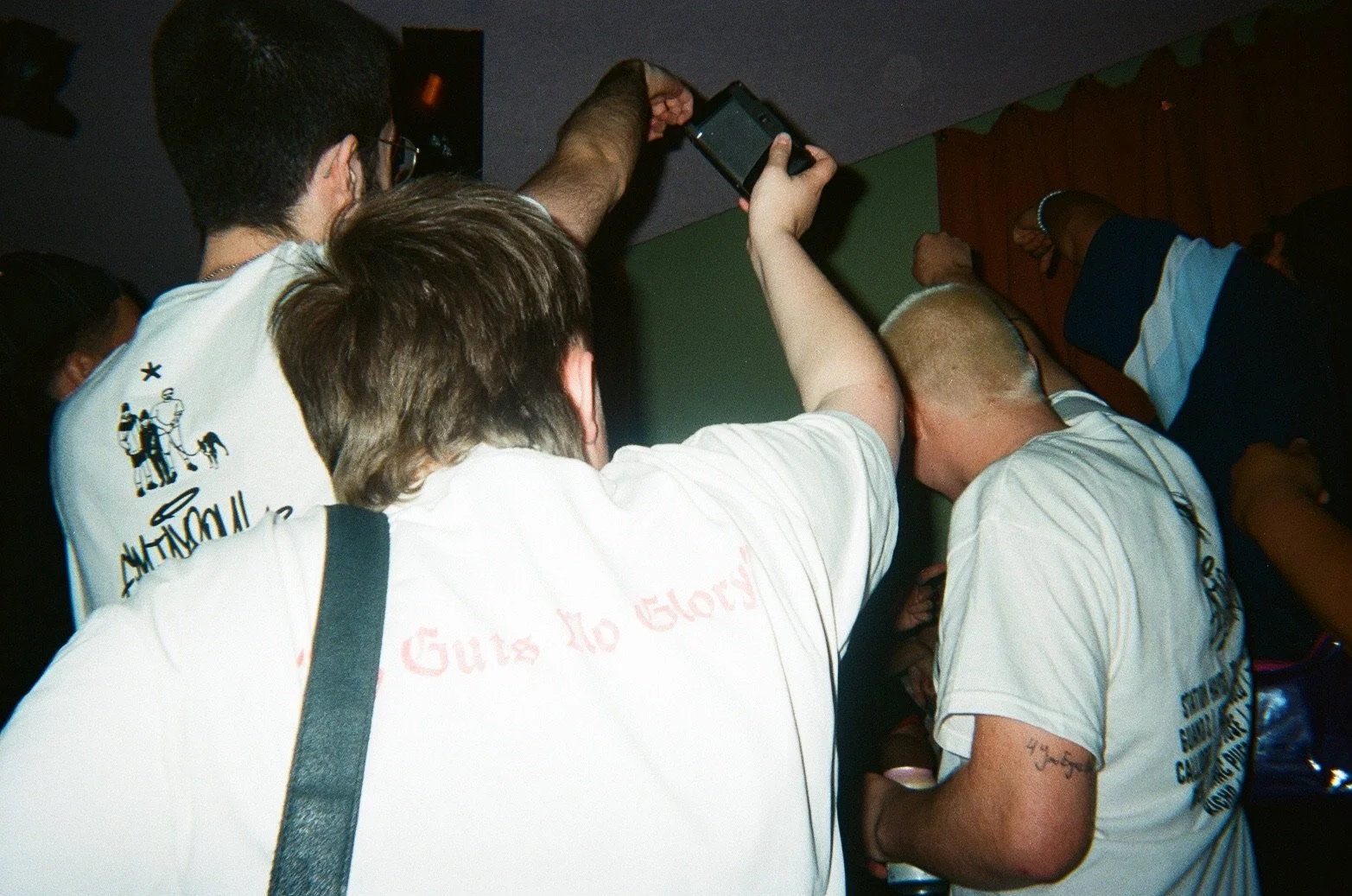 A group of young men gathered closely together, some appear to be taking a selfie with a smartphone, in a dimly lit indoor setting.