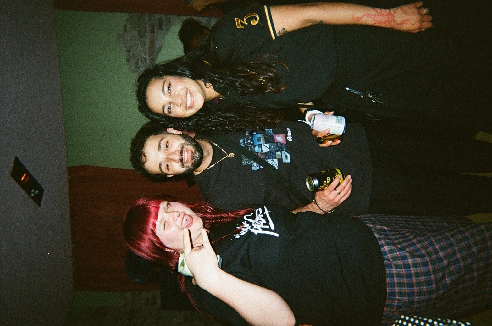 Three friends posing together at a party or social gathering, smiling and holding drinks, with a background featuring a green wall and a brick pattern.