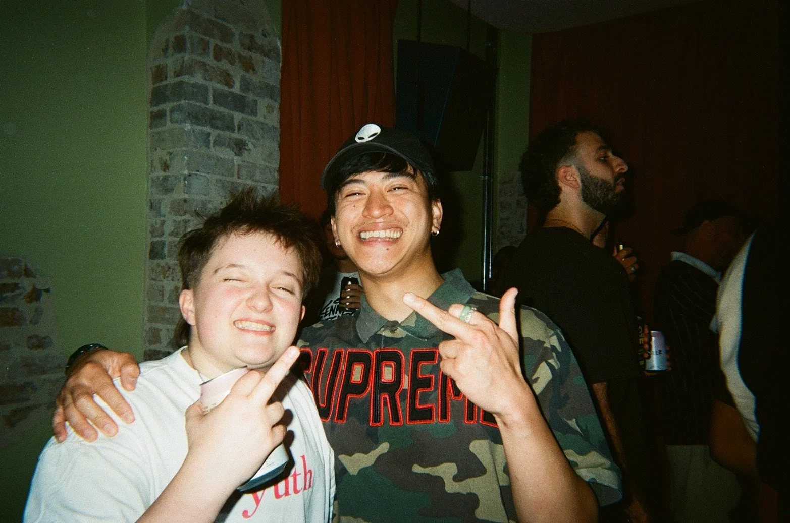Two young men smiling and making peace signs at a party, with other people in the background.