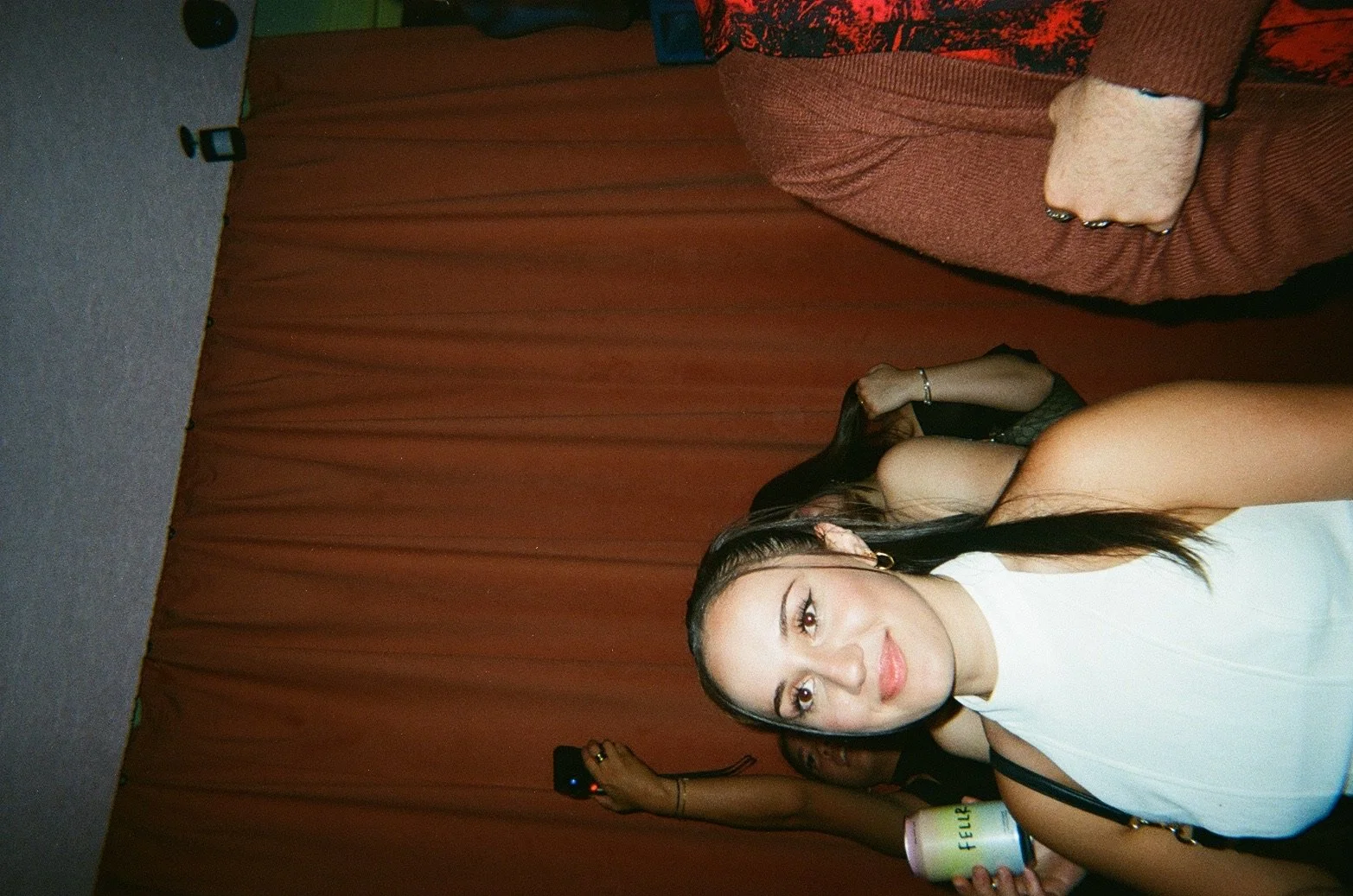 A young woman with long dark hair, wearing a white sleeveless top, posing indoors in front of orange-brown curtains, smiling slightly at the camera. There's a person in the background holding a cellphone.
