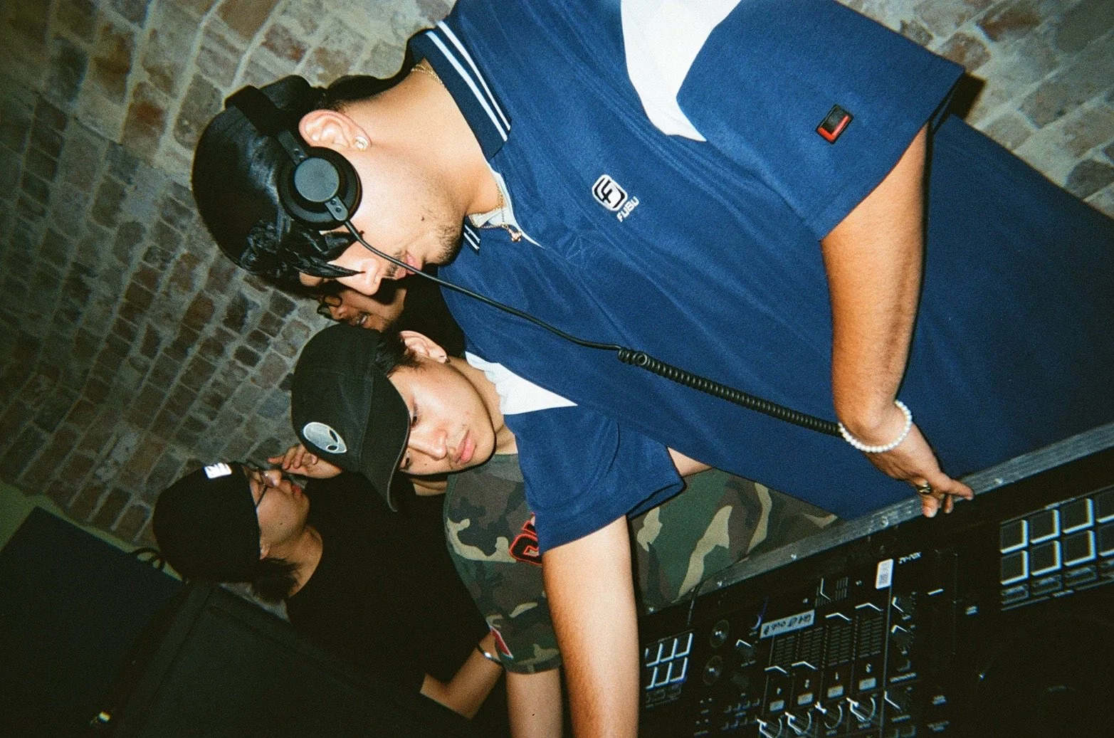 Three people DJing at a club with brick wall background, wearing headphones and caps, one in a blue outfit, another in black, and the third in camouflage shorts.