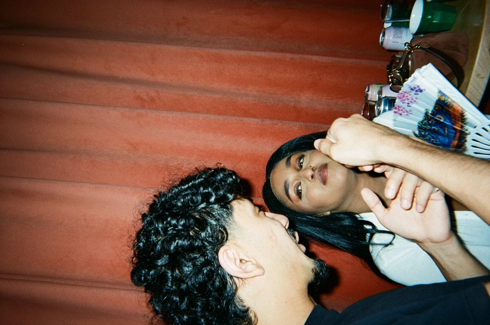 Two people close together, one with dark curly hair and the other with long dark straight hair, engaging in a conversation with the person on the right touching their face, against a background of a red curtain and a table with cans and a laptop.