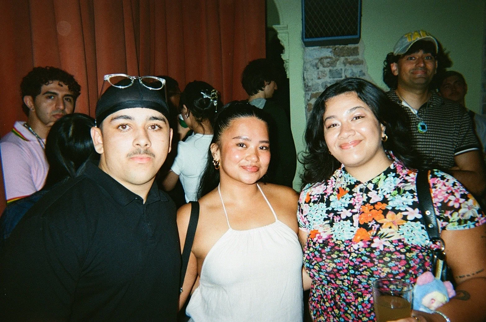 Group of diverse people at a party, three people in the foreground smiling, including a man with a mustache wearing a black shirt and head covering, a woman with dark hair in a white top, and a woman with black hair in a colorful floral dress holding