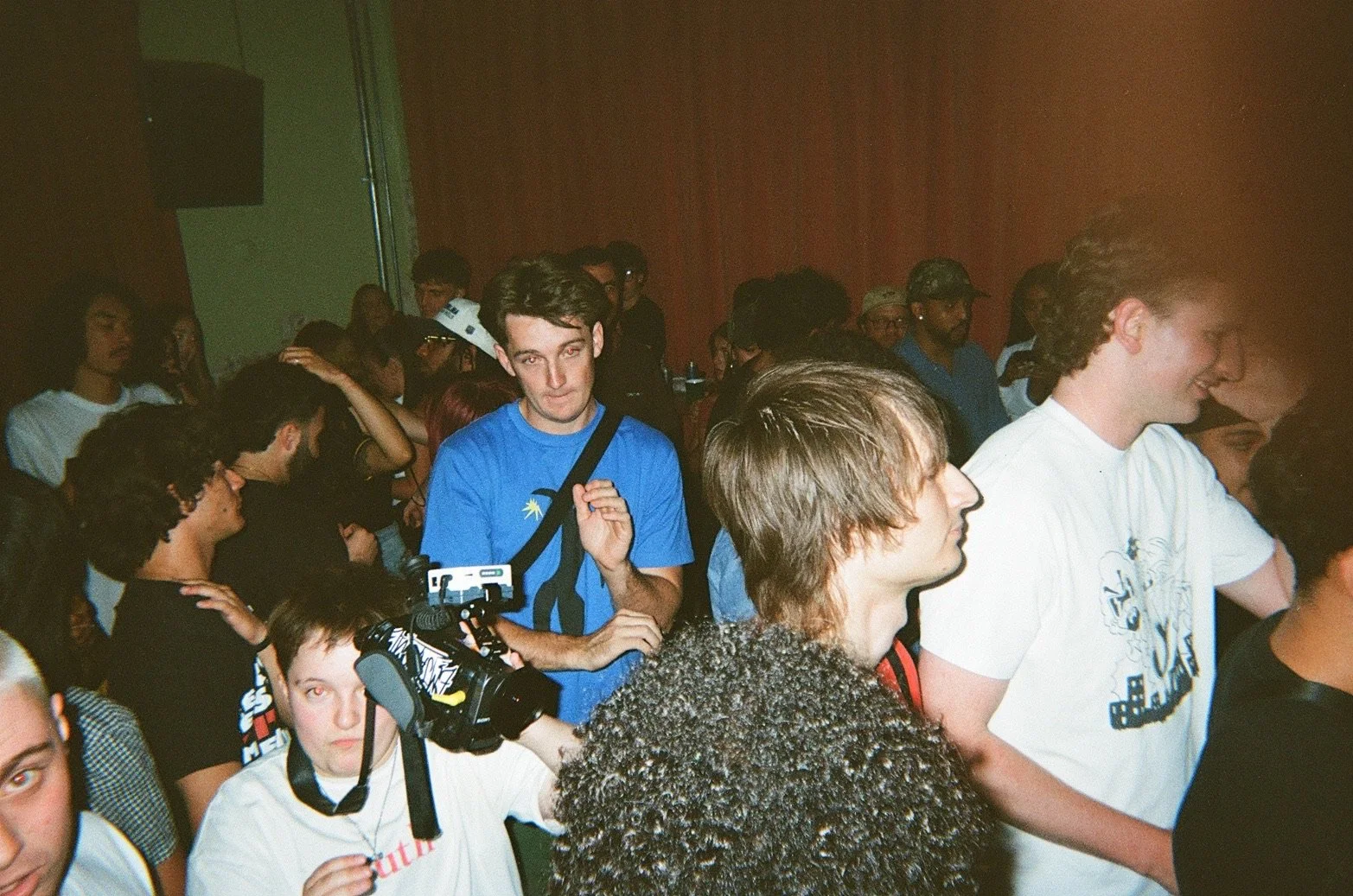 A crowded indoor event with several young people, some wearing t-shirts and hats, some with cameras, and others talking and socializing.