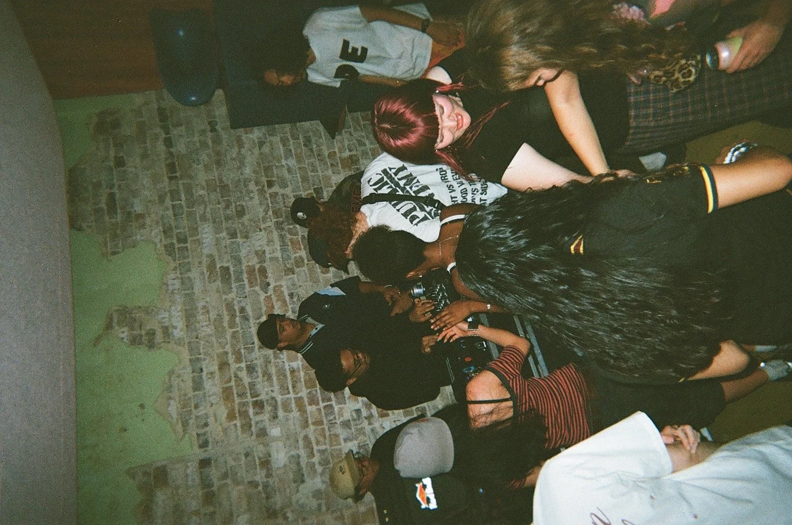Group of people standing close together in a bar or club with exposed brick wall, some wearing casual outfits and one with a camera.