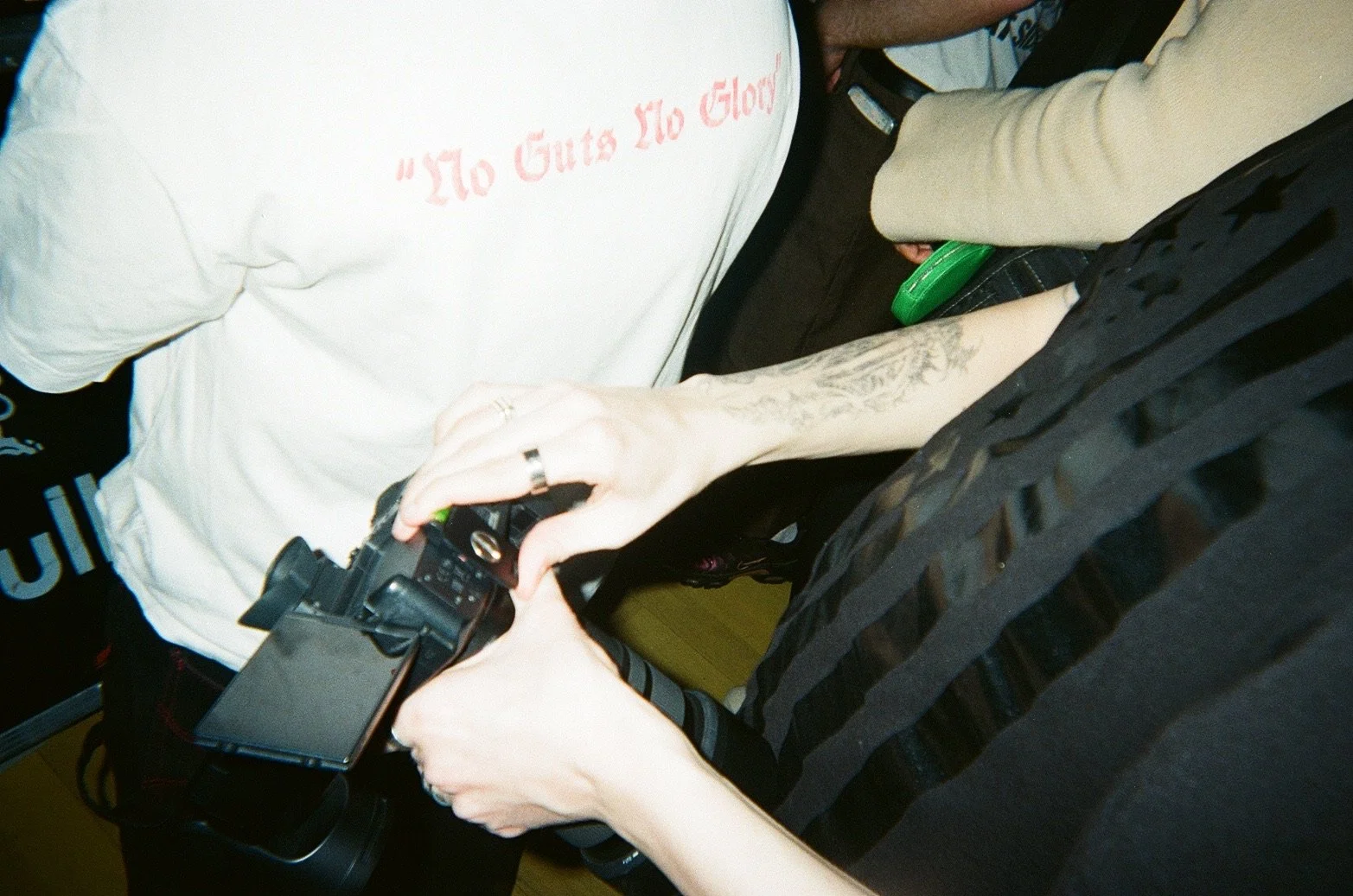 Close-up of a person holding a camera, with others around, one person wearing a white T-shirt with red text that reads 'To Guts To Glow'.