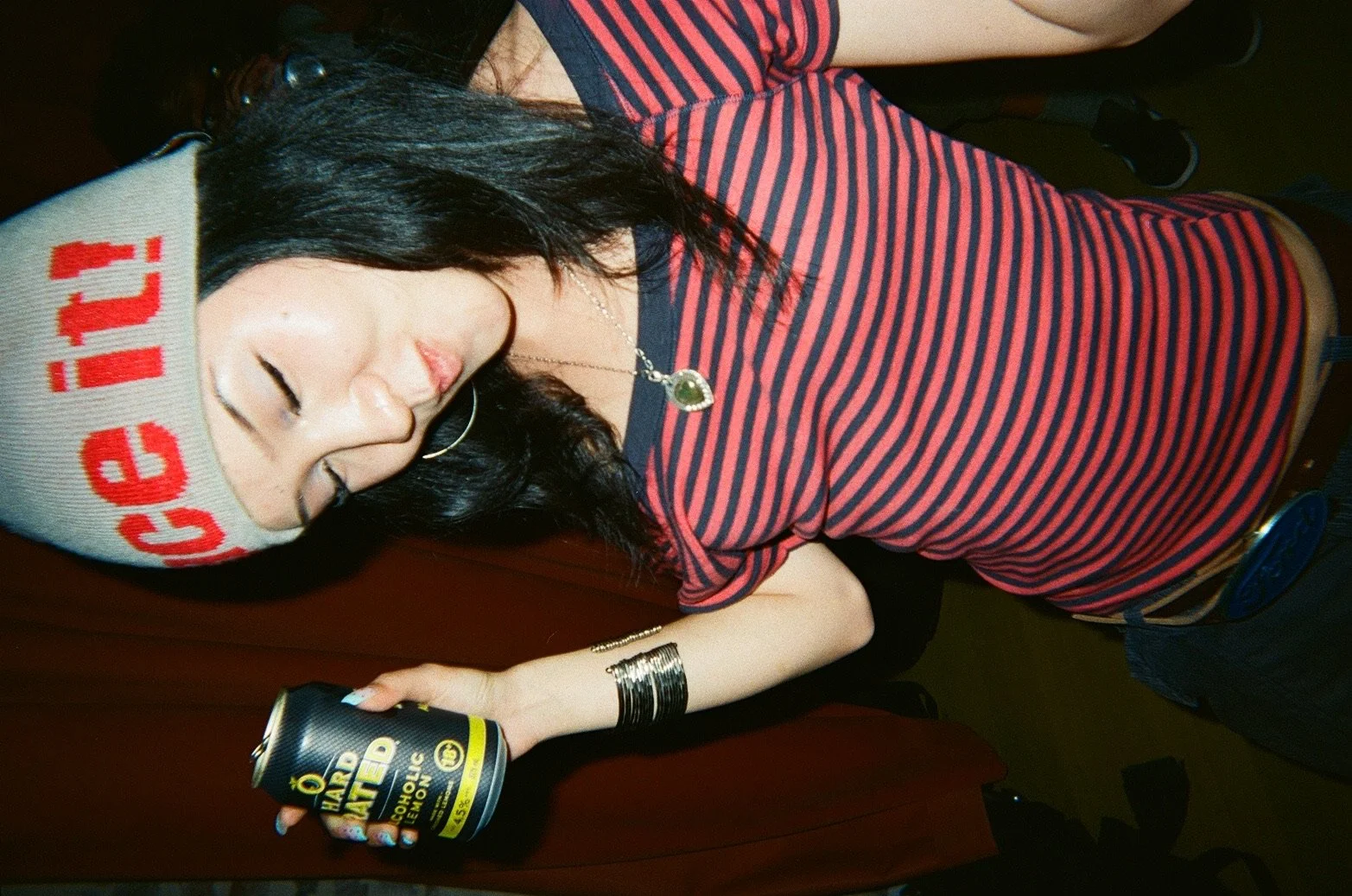 A young woman with black hair, wearing a white beanie with red writing, a red and navy striped shirt, and silver jewelry. She is holding a black and yellow can of alcoholic beverage and appears to be sleeping or resting with her eyes closed, lying on