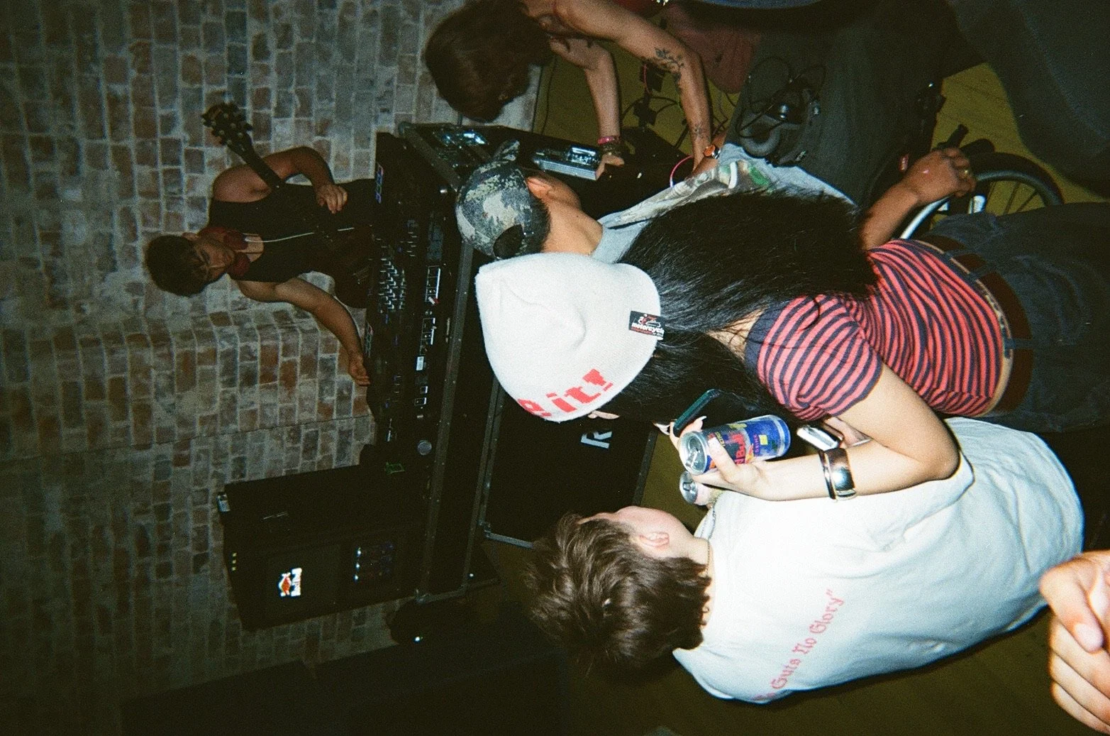 People gathered around a DJ setup in a dimly lit space with brick walls, some dancing and listening to music.