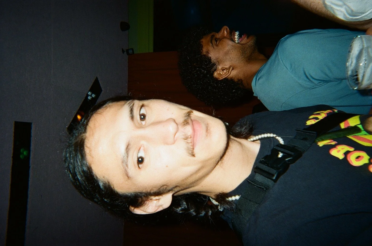 Two young men are laughing and smiling, one with a mustache and a black T-shirt, and the other with curly hair and a blue T-shirt, in an indoor setting.