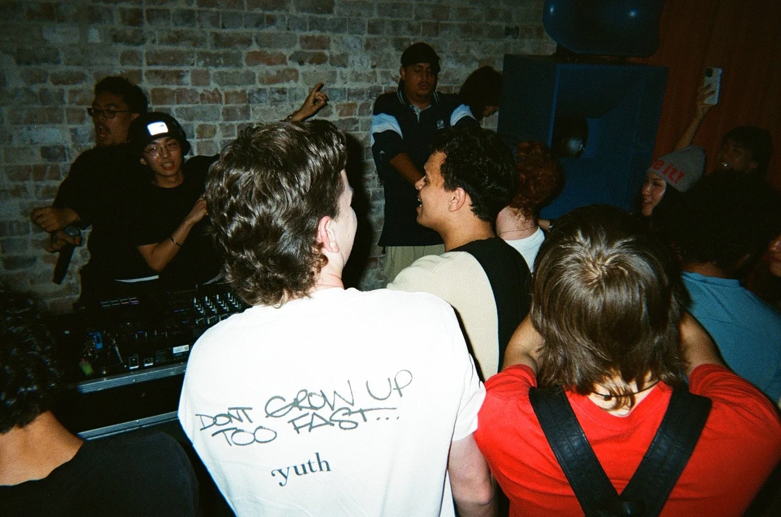A group of people gathered in a dimly lit room with a brick wall, likely at a social event or party. Some are wearing casual streetwear, and one person is wearing a gray beanie with red writing. There's a DJ setup in the foreground, and individuals a