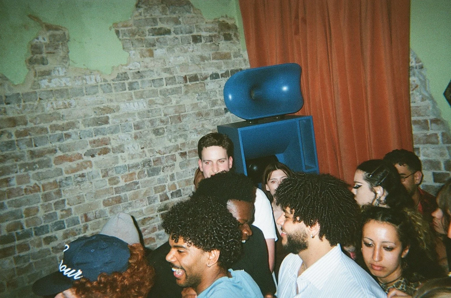 Group of diverse young people gathered indoors at a party, socializing and smiling, with exposed brick wall and a blue speaker in the background.