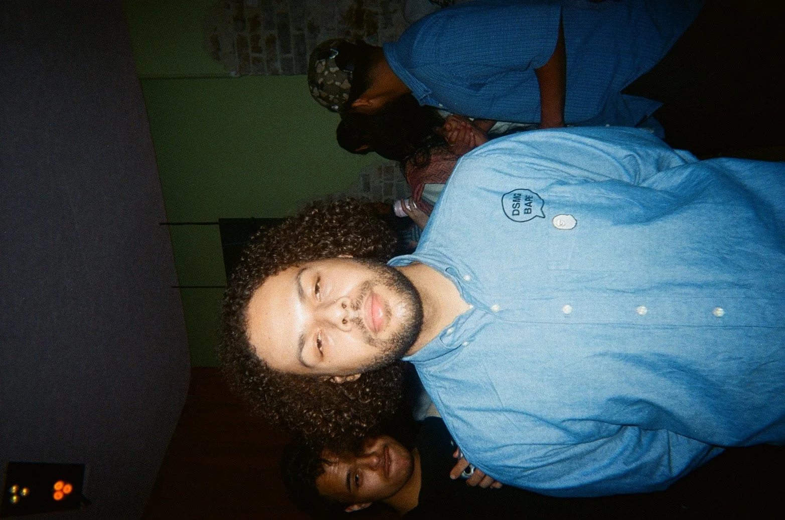 Young man with curly hair and a beard, wearing a light blue shirt, in a dimly lit club or bar, with other people in the background.