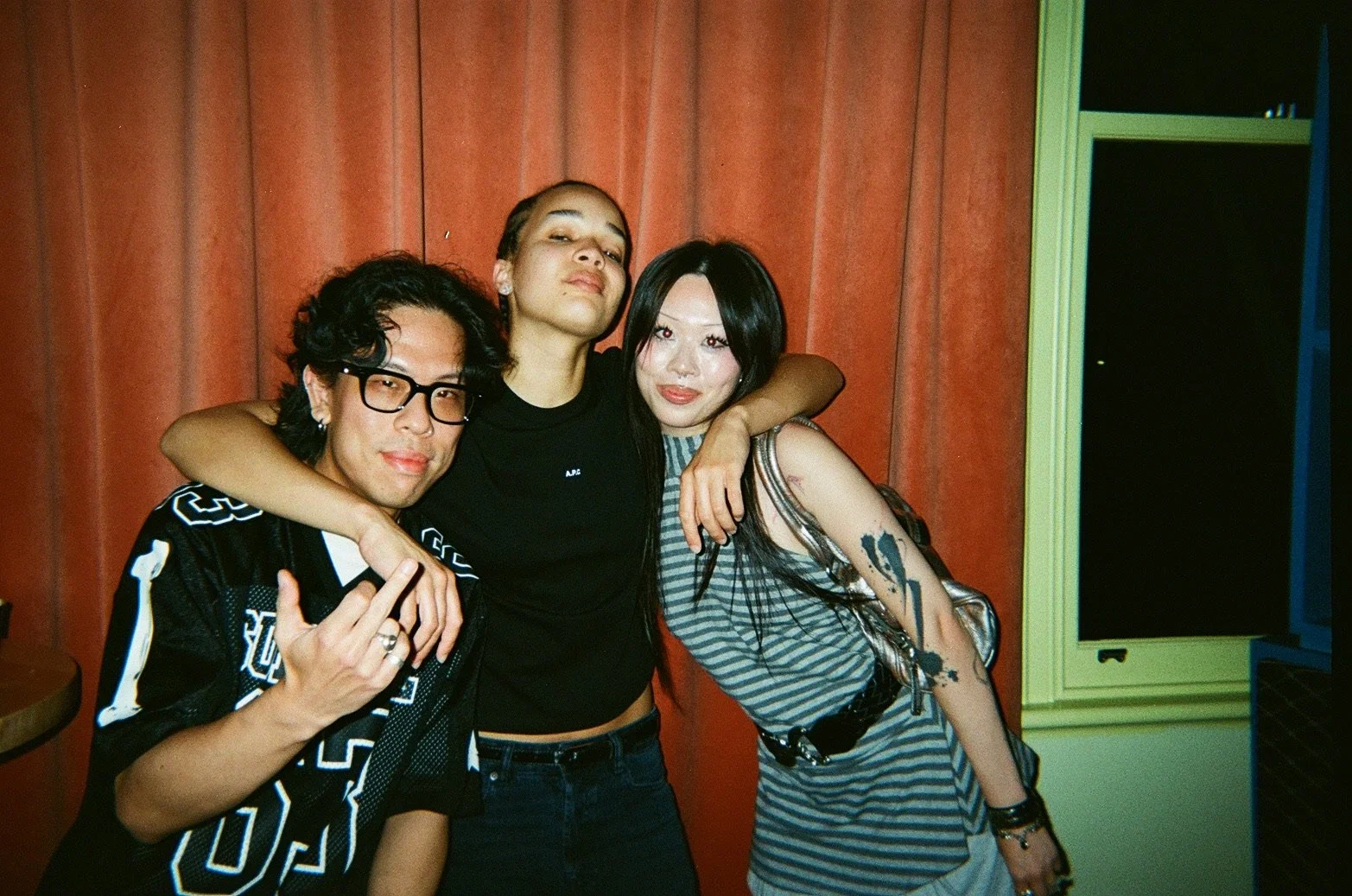 Three young adults pose together at a party, hugging in front of a red curtain with a yellow window frame to the right.