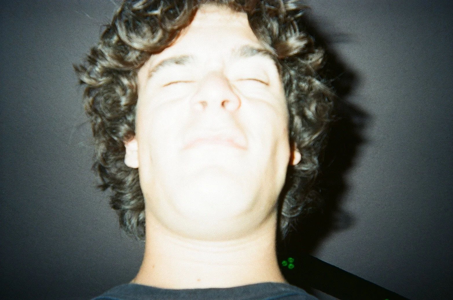 Close-up photo of a young person with curly hair and light skin, smiling with eyes closed, taken from a low angle against a dark background.